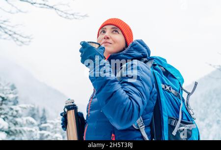 Frau mittleren Alters lächelt und trinkt ein heißes Getränk aus Thermoskanne gekleidet warme Daunenjacke, während sie Trekking Winter Berge Route. Aktive Peopl Stockfoto