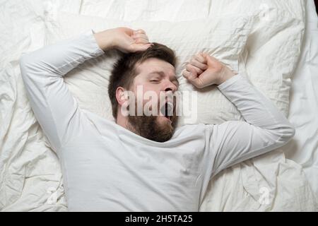 Niedlicher bärtiger Mann wacht aus dem Schlaf auf, er gähnt und streckt sich, während er im Bett liegt. Draufsicht. Nahaufnahme. Stockfoto