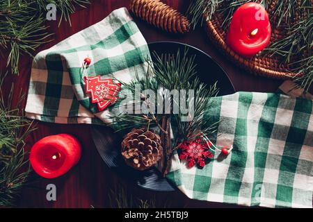 Festliche weihnachtliche Tischkulisse mit Tannenzweigen, roten Kerzen, Zapfen und Ornamenten. Schwarzer Teller mit grün karierter Serviette Stockfoto