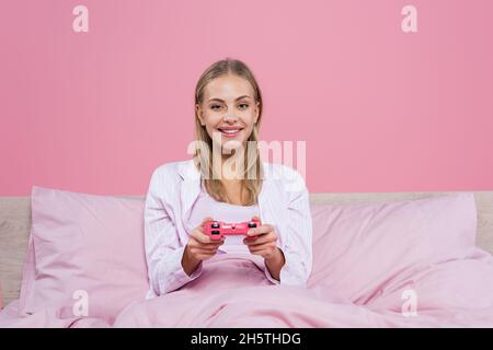 KIEW, UKRAINE - 27. OKTOBER 2020: Positive blonde Frau spielt Videospiel auf Bett isoliert auf rosa Stockfoto