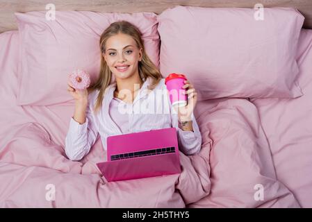 Draufsicht auf fröhliche Frau in Schlafanzug mit Papiertasse und Donut in der Nähe des Laptops auf dem Bett Stockfoto
