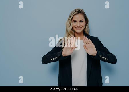 Lächelnde, höfliche junge Frau mit schwarzem Blazer über weißem T-Shirt, die die Hände in einer Stoppgeste hält Stockfoto