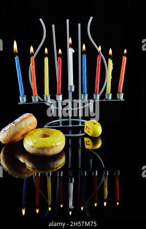 Happy Hanukkah und Hanukkah Sameach - traditioneller jüdischer Kerzenständer mit brennenden Kerzen und Donuts vor schwarzem Hintergrund mit Spiegelung. Stockfoto