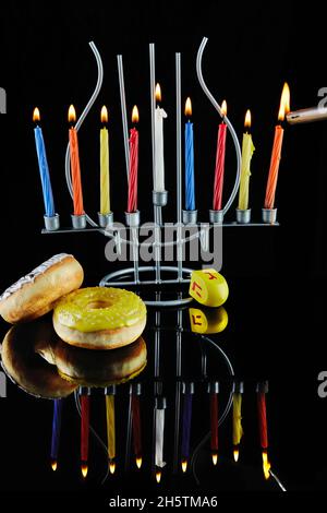 Happy Hanukkah und Hanukkah Sameach - traditioneller jüdischer Kerzenständer mit brennenden Kerzen und Donuts vor schwarzem Hintergrund mit Spiegelung. Stockfoto