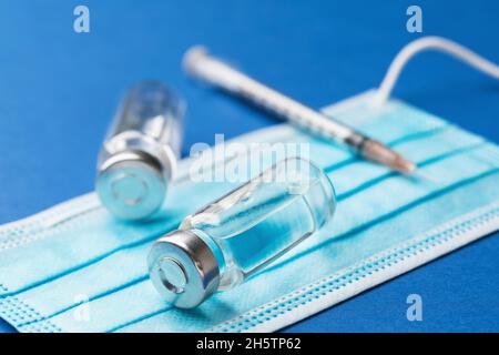 Zwei Fläschchen mit einer Injektionslösung, einer Einwegspritze und einer medizinischen Maske auf blauem Hintergrund, Impfkonzept Stockfoto