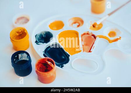 Auf dem weißen Tisch gibt es die Palette, die mit verschiedenen Farben der Farben gefärbt ist, die Gläser mit Gouache und den Pinsel. Der Prozess des Zeichnens und der Kreativität Stockfoto