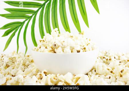 Popcorn in einer weißen Schüssel auf einem Hintergrund aus Palmblättern, auf einem weißen Teller. Selektiver Fokus. Mais-Snacks. Nahaufnahme. Stockfoto