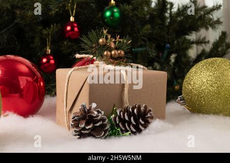 Schönes weihnachtsgeschenk mit bunten Kugeln auf Kunstschnee geschmückt, im Hintergrund ein weihnachtsbaum mit hängenden Glühbirnen, Feiertagsfeier Stockfoto