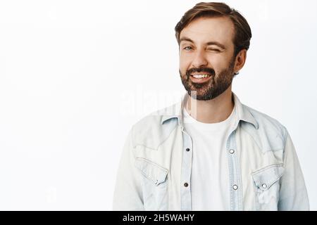 Charismatischer bärtiger Mann zwinkert und lächelt die Kamera an, flirtet und steht auf weißem Hintergrund Stockfoto
