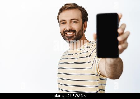 Porträt eines gutaussehenden lächelnden Casual Guy in T-Shirt, die Hand mit dem Telefon ausstreckend, zeigt Smartphone-Bildschirm, demonstriert App-Schnittstelle, weiß Stockfoto