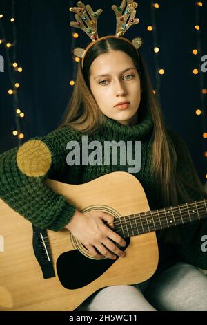 Ein schönes Teenager-Mädchen in weihnachtlichen Geweihen und ein grüner Pullover spielt Gitarre. Häusliche Umgebung, dunkler Hintergrund mit Bokeh aus Weihnachtslicht Stockfoto