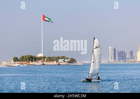 Abu Dhabi, VAE - 5. November 2021: Sonniger Tag in Abu Dhabi Corniche mit Segelboot im Vordergrund und Flagge der VAE im Hintergrund Stockfoto