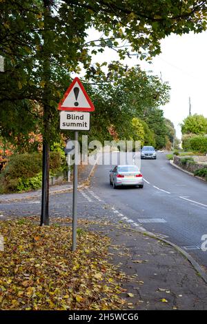 Ein Verkehrsschild in Großbritannien, das vor einem blinden Gipfel warnt, ist deutlich sichtbar. Es wird ein Fahrzeug gezeigt, das auf einer Wohnstraße zum blinden Gipfel fährt Stockfoto