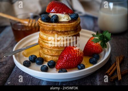Klare Sicht auf Stapel von Pfannkuchen mit Honigstock, Erdbeere, Blaubeere und Peitschencreme auf dem Teller, leckeres Dessert mit Milch zum Frühstück. Stockfoto