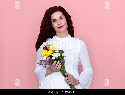 Frau hält Blumen mit weichem Lächeln auf rosa Hintergrund. Romantisches Porträt einer hübschen Frau in weißem Kleid mit Blumenstrauß über ihren Körper. Stockfoto