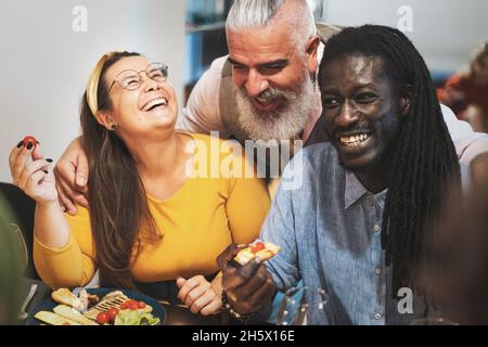Fröhliche interrassische Gruppe von erwachsenen Freunden, die zusammen essen, reden und scherzen - multirassische Gruppe von Erwachsenen, die am Esstisch sitzen Stockfoto
