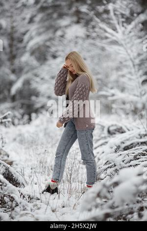 Eine niedliche junge Frau mit langen blonden Haaren in einer gestrickten, gemütlichen Strickjacke spaziert durch den winterlich verschneiten Wald Stockfoto