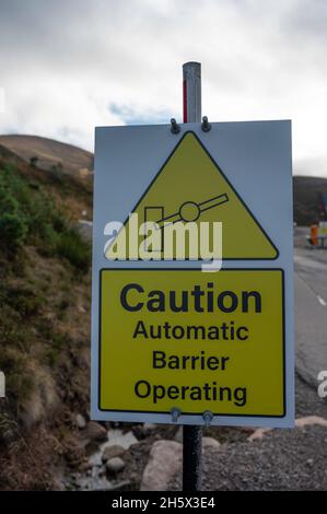 Gelbes und weißes Schild mit der Aufschrift „Achtung Automatische Barriere in Betrieb“. Barriere-Symbol. Verschwommener Hintergrund von Barriere, Straße und Bergen. Stockfoto