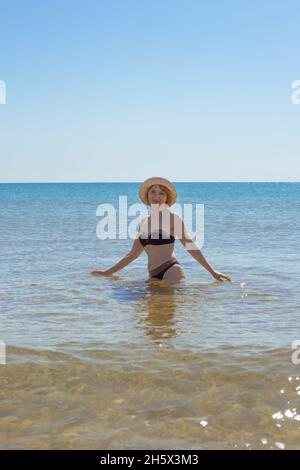 Europäische Frau im Badeanzug und hüftlangen Hut im Meer lächelt und schaut in die Kamera. Urlaub am Meer. Stockfoto