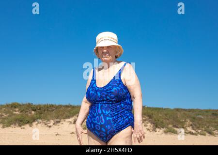 Eine ältere Frau in einem blauen Badeanzug und Hut steht am Meer vor dem Hintergrund eines blauen Himmels. Ruhe auf dem Meer im Ruhestand. Stockfoto