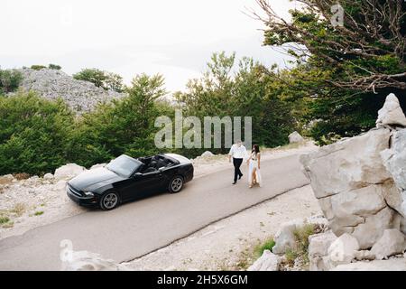 Die Braut und der Bräutigam gehen mit Händen in der Nähe eines offenen Autos auf der malerischen Straße in den Bergen in Montenegro Stockfoto