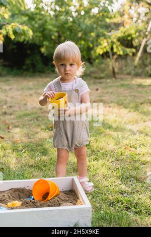 Ein kleines Mädchen in einem grauen Jumpsuit mit Shorts und einem gelben Spielzeugeimer mit Sand in den Händen spielt im Garten und schaut auf die Kamera. Dort Stockfoto