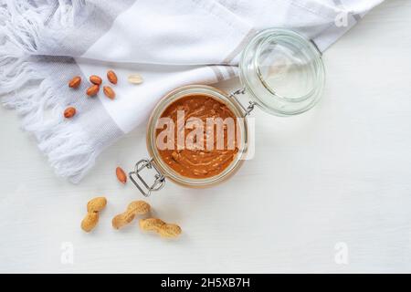 Erdnussbutter im Glas auf weißem Holztisch mit Geschirrtuch. Draufsicht, flach liegend, Kopierbereich Stockfoto