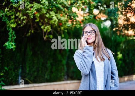 Positive Arbeitnehmerin mit fadem Haar, die auf dem Bürgersteig steht und auf dem Handy spricht, während sie wegschaut Stockfoto