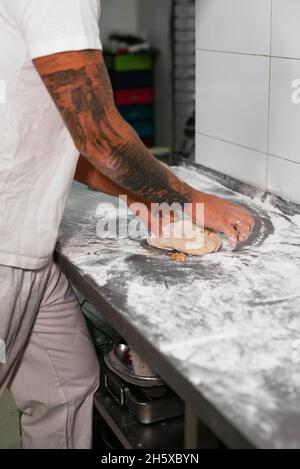 Seitenansicht des tätowierten männlichen Bäckerers in weißem Polohemd, der Teig mit den Händen knetet, während er an der Metalltheke in der Küche der modernen Bäckerei steht Stockfoto