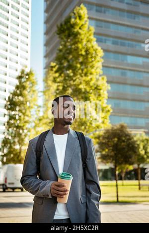Positiver afroamerikanischer Arbeiter mit Rucksack, der mit einer Einweg-Tasse Kaffee steht und mit toothy Lächeln wegschaut Stockfoto
