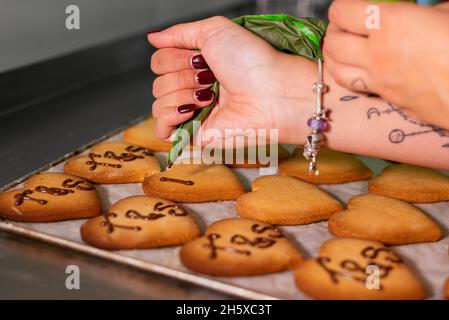 Crop junge weibliche Konditorin Dekoration herzförmigen Zucker Cookies mit Schokolade Glasur stehen am Tisch in der Bäckerei Stockfoto