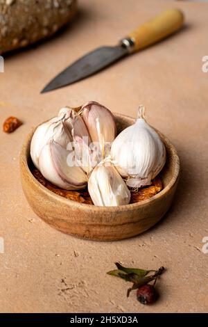 Nelken von ungeschältem Knoblauch mit weißer Schale, serviert in einer kleinen runden Holzschüssel, die auf einem braunen Tisch in der hellen Küche aufgestellt wurde Stockfoto