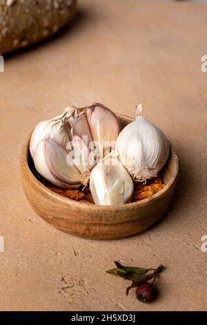 Nelken von ungeschältem Knoblauch mit weißer Schale, serviert in einer kleinen runden Holzschüssel, die auf einem braunen Tisch in der hellen Küche aufgestellt wurde Stockfoto