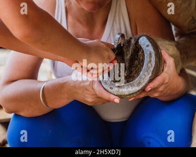 Crop unkenntlich Horsewoman mit Assistent Reinigung Schlamm aus Hufeisen mit Hufeisen in Stall Stockfoto