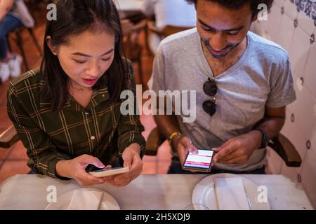 Glückliches junges multirassisches Paar in legerer Kleidung mit Smartphones, während es zusammen am Tisch im modernen Restaurant sitzt Stockfoto