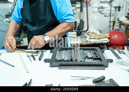 Hoher Erntewinkel unkenntlich älterer männlicher Handwerker in Schürze und Brille, der Metallrahmen für den Buchdruck im Studio vorbereitet Stockfoto