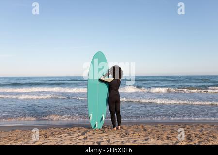 Rückansicht einer nicht erkennbaren weiblichen Surferin im Neoprenanzug mit Surfbrett, das auf die vom Meer geschwemmte Küste blickt Stockfoto