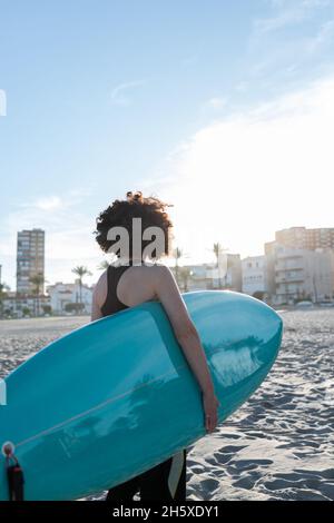 Rückansicht einer nicht erkennbaren weiblichen Surferin im Neoprenanzug mit Surfbrett, das auf die vom Meer geschwemmte Küste blickt Stockfoto
