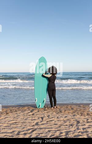 Rückansicht einer nicht erkennbaren weiblichen Surferin im Neoprenanzug mit Surfbrett, das auf die vom Meer geschwemmte Küste blickt Stockfoto