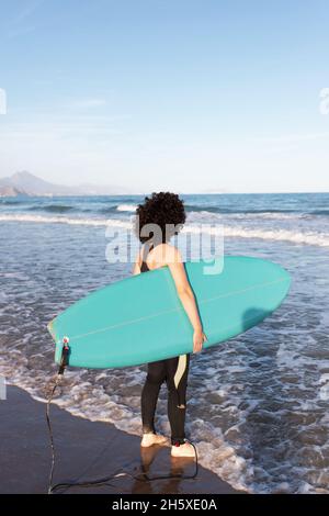 Rückansicht einer nicht erkennbaren weiblichen Surferin im Neoprenanzug mit Surfbrett, das auf die vom Meer geschwemmte Küste blickt Stockfoto