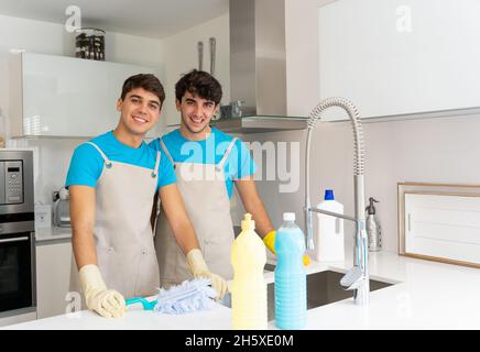 Positive junge männliche Putzfrauen in Uniformen und Gummihandschuhen lächeln und blicken auf die Kamera, während sie bei Tageslicht in der modernen Küche arbeiten Stockfoto