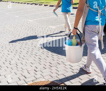 Unbekennbare junge Putzkolonne in Uniform, die Eimer trug, während sie an sonnigen Tagen auf einer gepflasterten Straße in der Nähe von Wohnhäusern ging Stockfoto