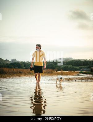 Ganzer Körper des männlichen Besitzers mit Stiefeln in den Händen zu Fuß im Wasser in der Nähe Laufhund am Sommertag in der Natur Stockfoto