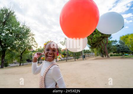 Fröhliche afroamerikanische Mädchen mit Zöpfen in stilvoller Kleidung läuft mit bunten Ballons in der Hand im Park tagsüber Stockfoto