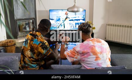 Von oben Rückansicht von anonymen multirassischen männlichen Freunden, die auf der Couch mit Gamepads in den Händen sitzen und Videospiele im Wohnzimmer spielen Stockfoto