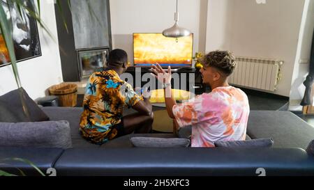 Von oben Rückansicht von anonymen multirassischen männlichen Freunden, die auf der Couch mit Gamepads in den Händen sitzen und Videospiele im Wohnzimmer spielen Stockfoto