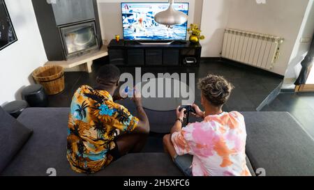Von oben Rückansicht von anonymen multirassischen männlichen Freunden, die auf der Couch mit Gamepads in den Händen sitzen und Videospiele im Wohnzimmer spielen Stockfoto