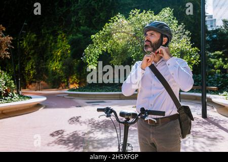 Erwachsener bärtiger männlicher Manager in formeller Kleidung, der den Helm für Fahrradfahren im Stadtpark anpasst Stockfoto