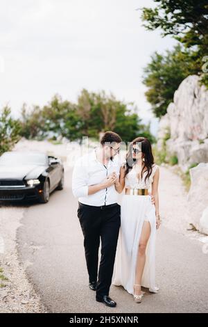 Die Braut und der Bräutigam gehen mit Händen in der Nähe eines offenen Autos auf der malerischen Straße in den Bergen in Montenegro Stockfoto