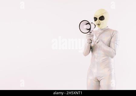 Person in silbernem Anzug und grüner Alien-Maske gekleidet, spricht durch ein weißes Megaphon auf weißem Hintergrund Stockfoto
